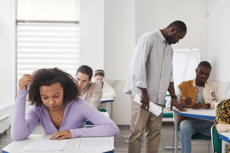 Teenage student at a classroom desk with her head down and a hand in her hair while a teacher stands over students behind her