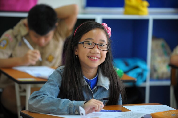 Smiling elementary student in glasses seated at her classroom desk, calm and ready to work, with a classmate writing behind her