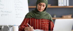 Teacher writing notes in a notebook at her desk beside a laptop